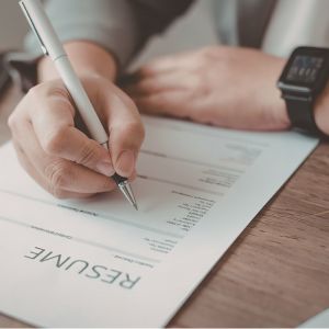 Close-up of a hand signing a resume on a desk, with a pen and a smartwatch visible on the wrist