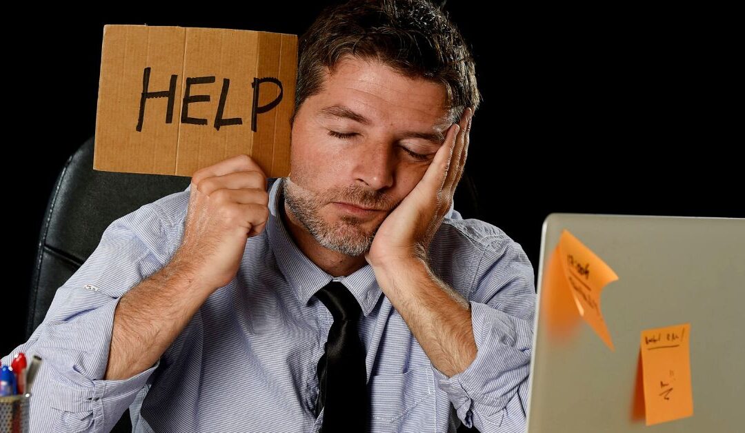 man with eyes closed sitting in front of lap top holding a cardboard help sign