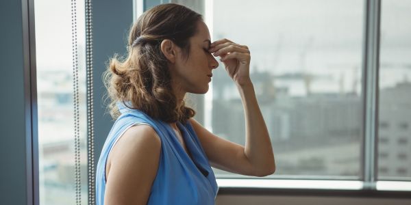 Woman in a blue sleeveless top sits by a window, pinching the bridge of her nose in apparent headache.