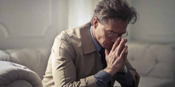 Middle-aged man wearing a beige jacket sits on a couch with hands pressed together in prayer.