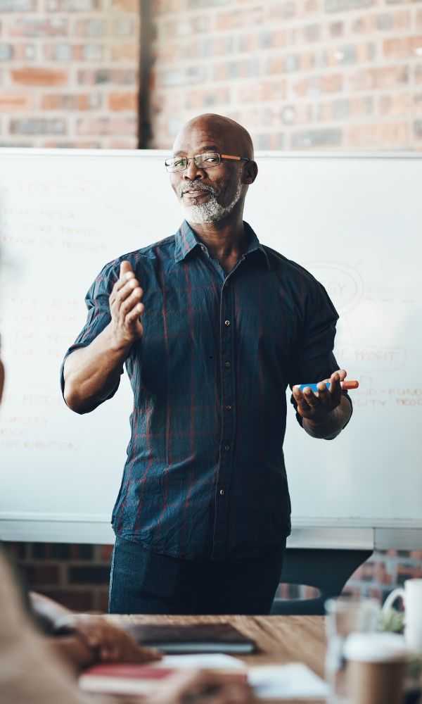 African American man with glasses giving a presentation in front of a whiteboard, gesturing with his hands while speaking.