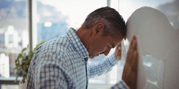 Man in a blue checkered shirt rests his forehead on a glass wall, looking down in a contemplative moment in an office setting.