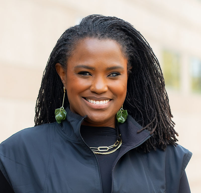Smiling Black woman with long dreadlocks wearing a dark jacket and green leaf-shaped earrings, outdoors with a blurred background.