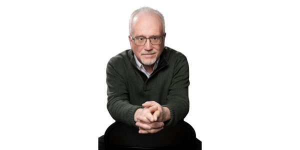 Portrait of an older man with glasses, gray hair, wearing a dark green sweater, hands clasped, facing the camera against a white background.